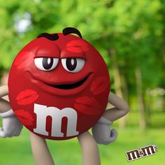 a red m & m's ball with eyes and hands on its back, sitting in the grass