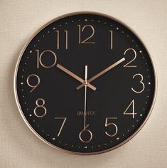 a black and gold clock with numbers on the face is hanging from a wall in front of a beige background