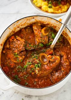 a pot filled with meat and vegetables on top of a white countertop next to a wooden spoon