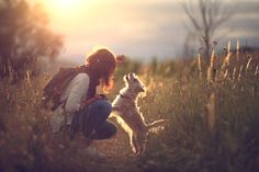 a woman kneeling down next to a dog on top of a grass covered field at sunset