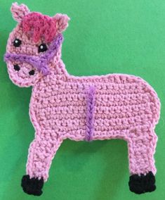 a crocheted pink horse with a heart on its nose and tail, standing against a green background