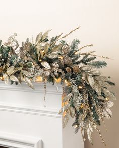 a white fireplace mantel decorated with gold and silver christmas garland, lights and greenery