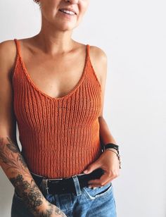 a woman in an orange tank top with tattoos on her arm and arms crossed, looking at the camera