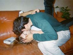 a man and woman cuddle on the back of a brown leather couch in front of a potted plant