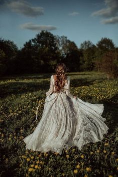 a woman in a long white dress walking through a field full of dandelions