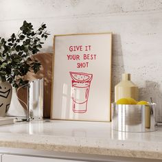 a framed poster that says, give it your best shot on a kitchen counter next to two vases and a potted plant