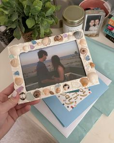 a person holding up a photo frame with seashells and shells on it next to a potted plant