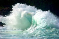 a large wave crashing into the ocean with green water and rocks in the back ground