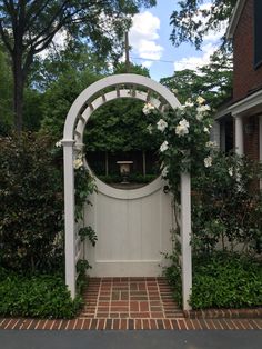 a white gate surrounded by greenery with flowers on the arch and brick walkway leading up to it