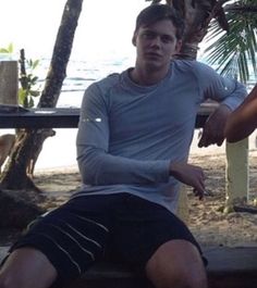 a man sitting on a bench next to another man near the water and palm trees