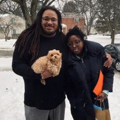 two people standing in the snow with a dog on their lap and one person holding a bag