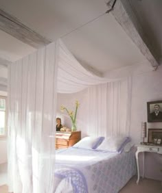 a white bed sitting in a bedroom next to a wooden dresser and window with sheer curtains