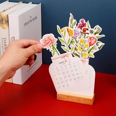 a hand holding a calendar with flowers in it on top of a table next to books