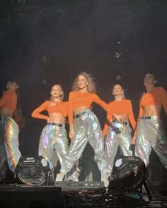 the dancers are wearing shiny silver pants and orange tops, while they stand on stage with their hands in their pockets