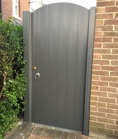 an open grey gate in front of a brick wall and shrubbery on the sidewalk