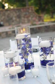 blue flowers and candles are in glass vases on a white table cloth with water