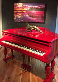 a red piano in front of a painting on the wall and wooden floor with an animal figurine sitting on top