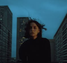 a woman standing in front of tall buildings at night with her hair blowing in the wind