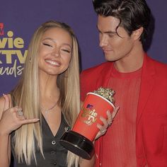 a man holding a popcorn cup next to a blonde woman in a black dress and red blazer