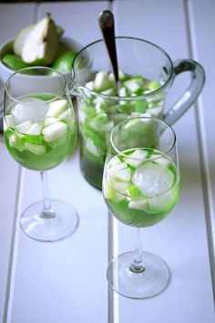 two wine glasses filled with green liquid and sliced apples in the background on a white table