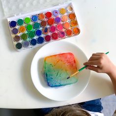 a person is painting with watercolors on a white plate next to a paintbrush