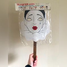 a person holding up a paper mask on top of a wooden stick in front of a white wall