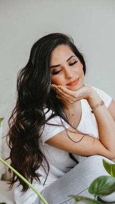 a woman with long dark hair sitting on a white couch next to a green plant