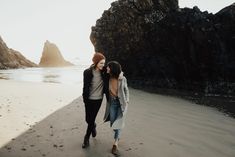 two young women walking on the beach next to some rocks and water, one is wearing a red beanie