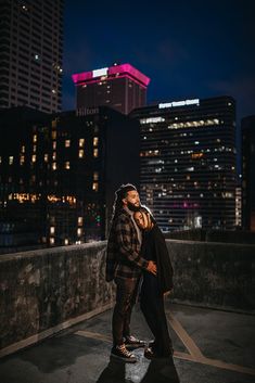 a man and woman standing on top of a roof at night with city lights in the background