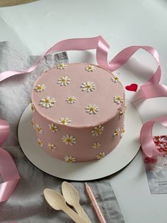 a pink cake decorated with daisies on a plate next to ribbon and spoons