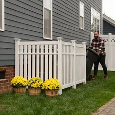 a man holding a wheelbarrow next to a white picket fence with yellow flowers