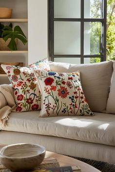 a white couch with pillows on it in front of a window and a bowl sitting on the coffee table