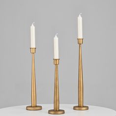 three brass candlesticks on a white table with one candle in the middle and two standing upright