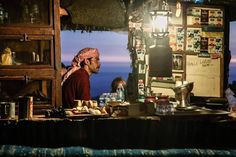 a man standing in front of a counter with food on it and people sitting around