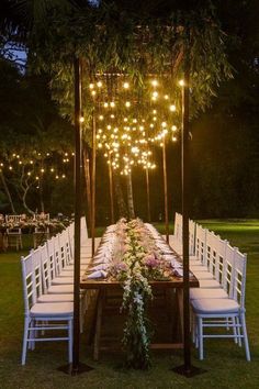 an outdoor dinner table with white chairs and lights hanging from the ceiling over it, surrounded by greenery