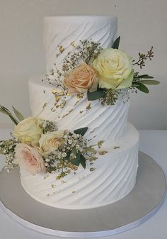 a three tiered white wedding cake with flowers on the top and sides, sitting on a silver platter