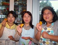 three women standing in front of a window holding up pastries to the camera while one woman holds out her hand