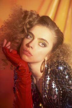 a woman with long curly hair and red gloves posing for a photo in front of an orange background