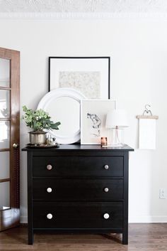 a dresser with a mirror, lamp and pictures on it in a white walled room