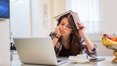 a woman sitting at a table with a laptop and book on top of her head