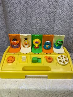 an assortment of sesame street toys sitting in a yellow plastic box on a white tablecloth