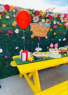 a yellow table topped with cakes and desserts next to a wall covered in flowers