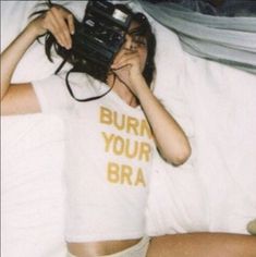 a woman laying in bed while holding a camera to her face with the words burn your bra on it