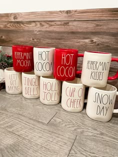 six coffee mugs with words on them sitting next to a christmas tree in front of a wooden wall