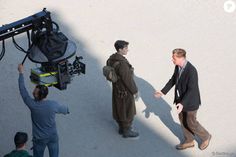 two men standing next to each other in front of a camera and another man pointing at something