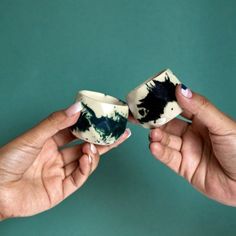 two hands are holding small cups with black and white designs on them, against a teal green background