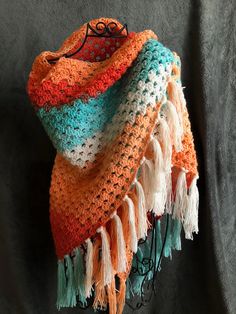 an orange, white and blue crocheted scarf hanging on a black wall with grey fabric