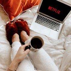 a person sitting on a bed holding a cup of coffee in front of a laptop