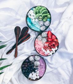 three bowls filled with different types of food on top of a white blanket next to two spoons