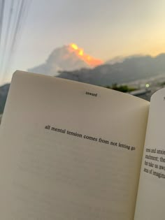 an open book sitting on top of a wooden table next to a window with mountains in the background